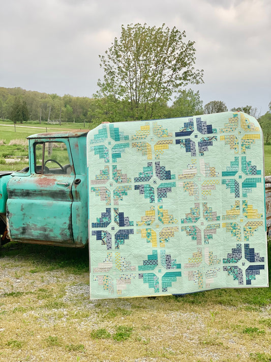 Colorful quilt featuring geometric patterns in mint green, navy, yellow, and teal, displayed on a vintage truck.