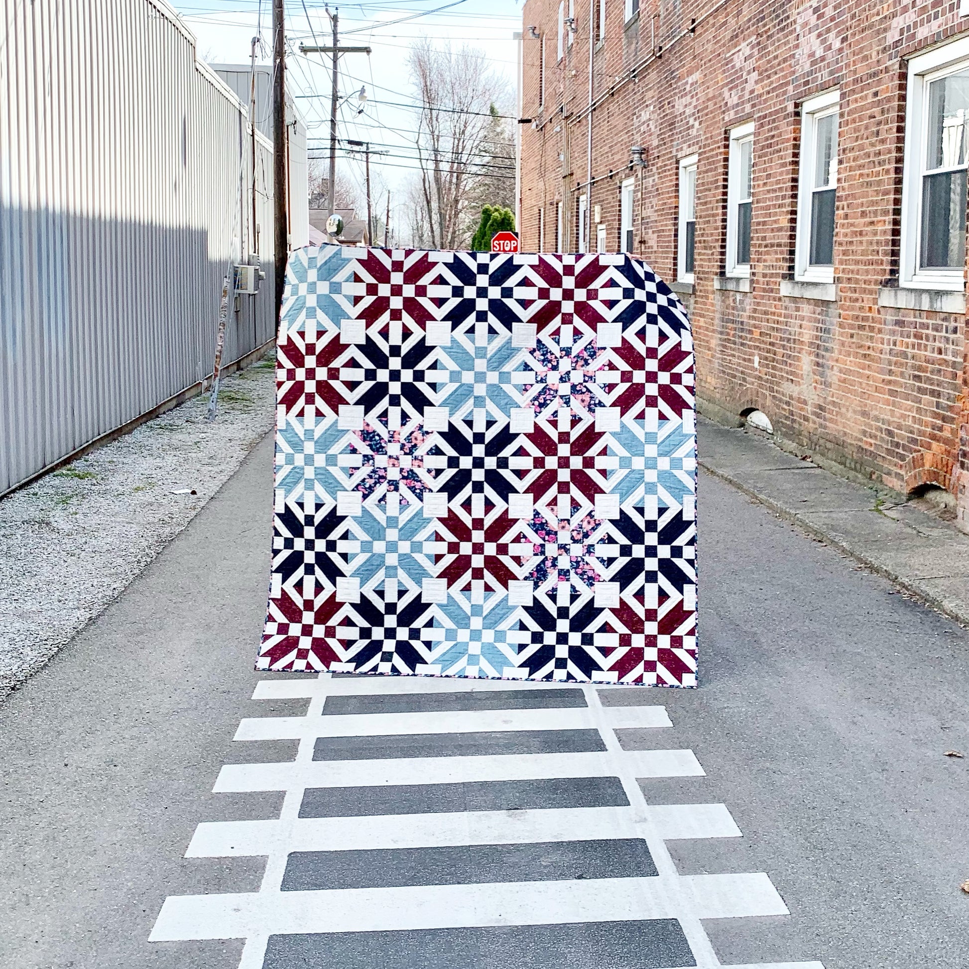 Colorful quilt featuring geometric patterns in red, white, and blue, displayed in an urban alley.