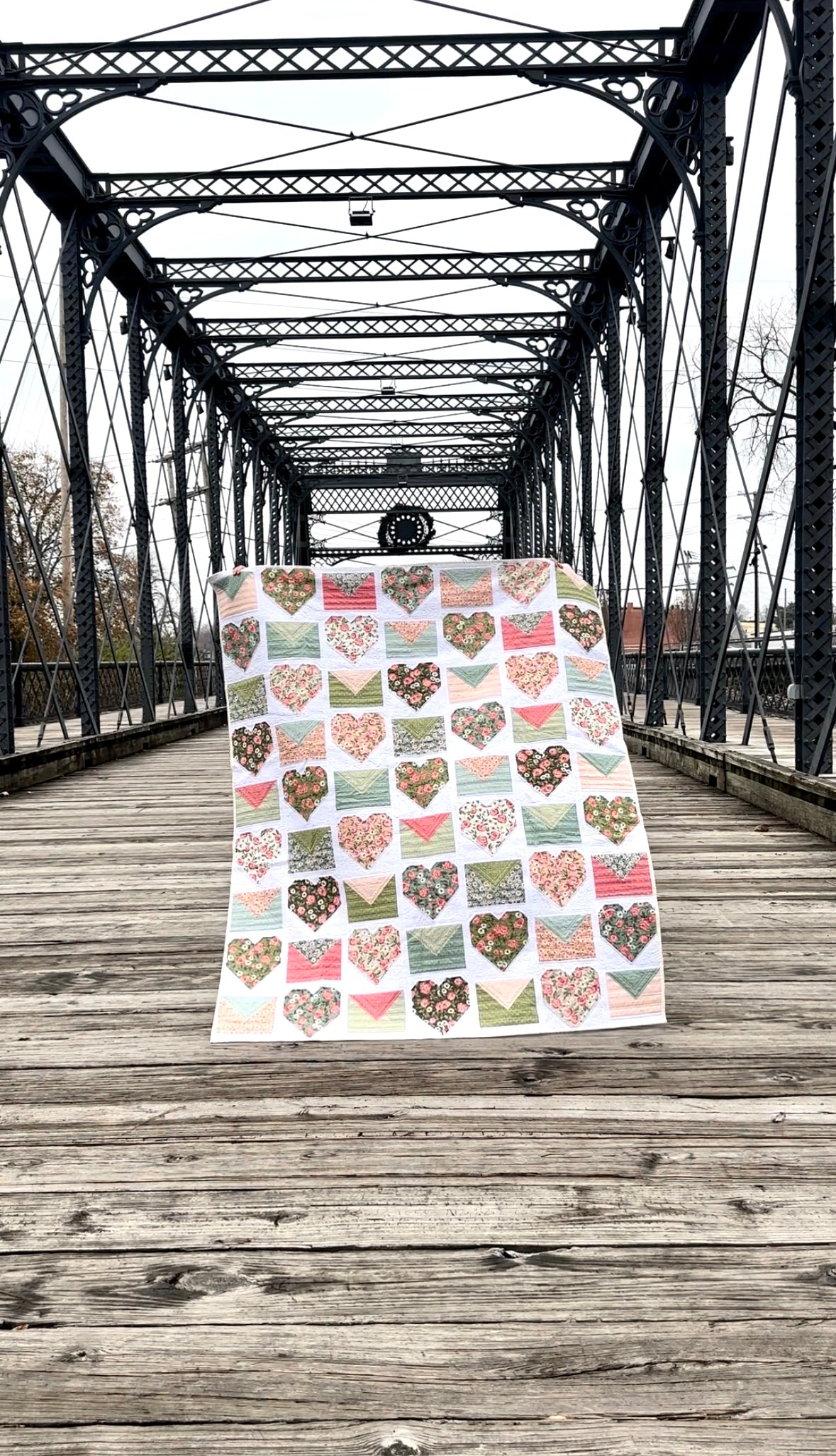 Colorful heart-patterned quilt, featuring floral designs in pink, green, and white, displayed on a rustic wooden bridge.