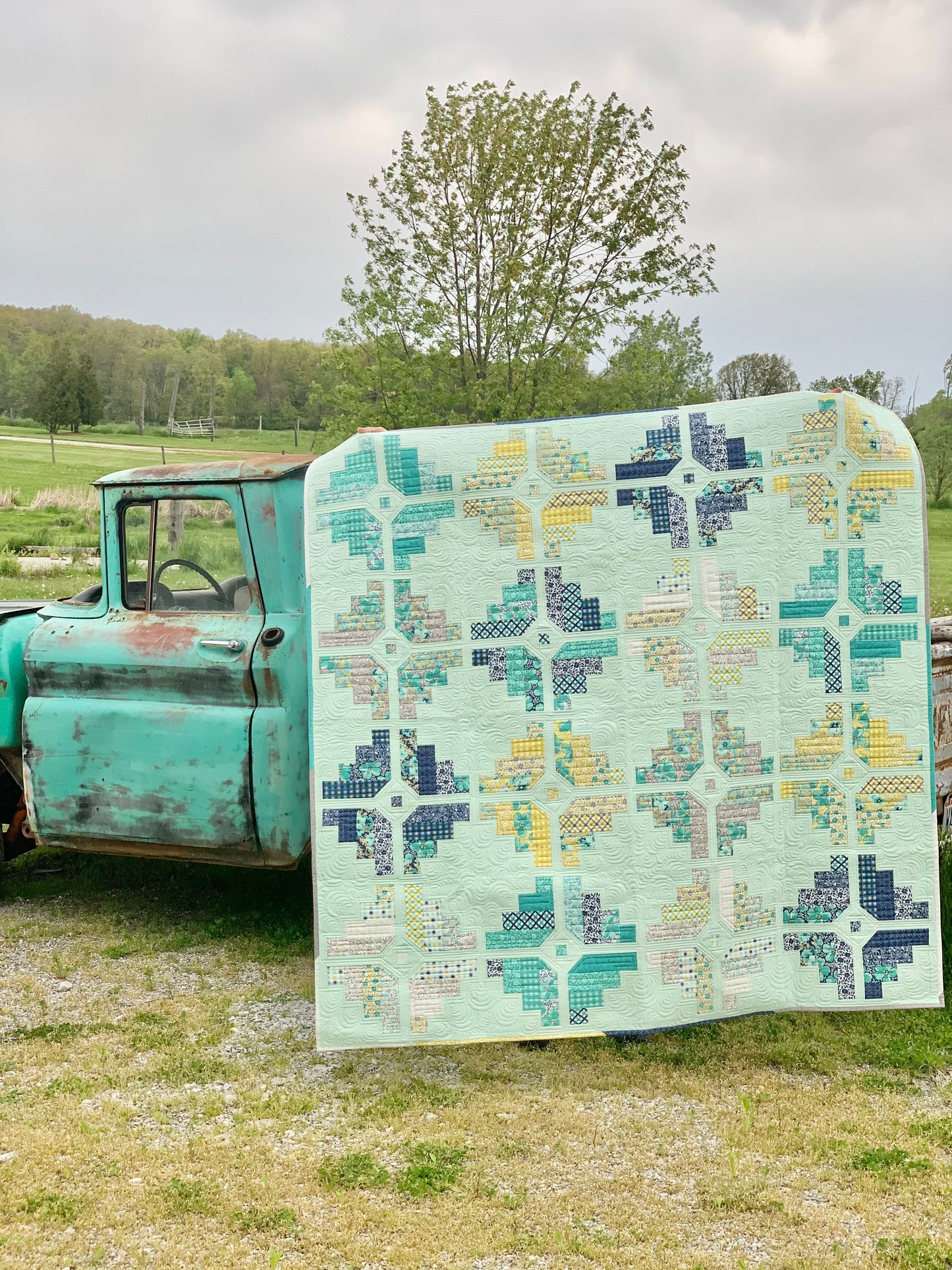 Colorful quilt featuring geometric patterns in mint green, navy, yellow, and teal, displayed on a vintage truck.
