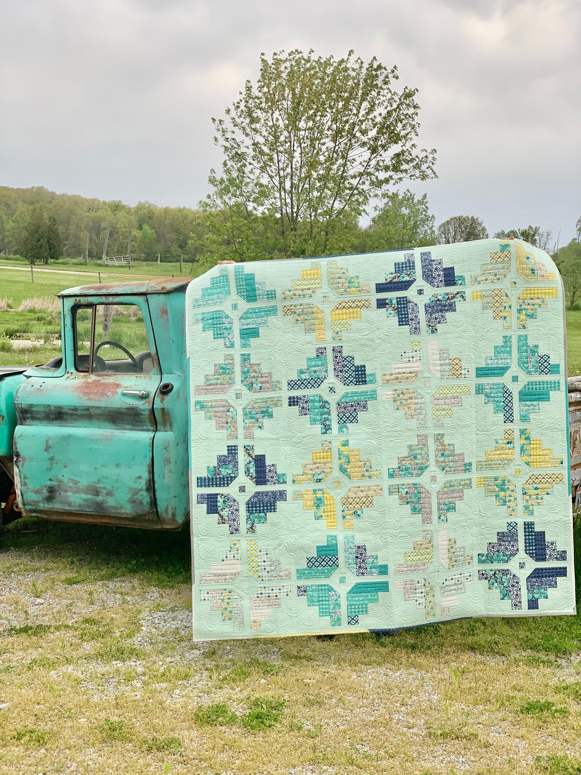 Colorful quilt featuring geometric patterns in mint green, navy, yellow, and teal, displayed on a vintage truck.