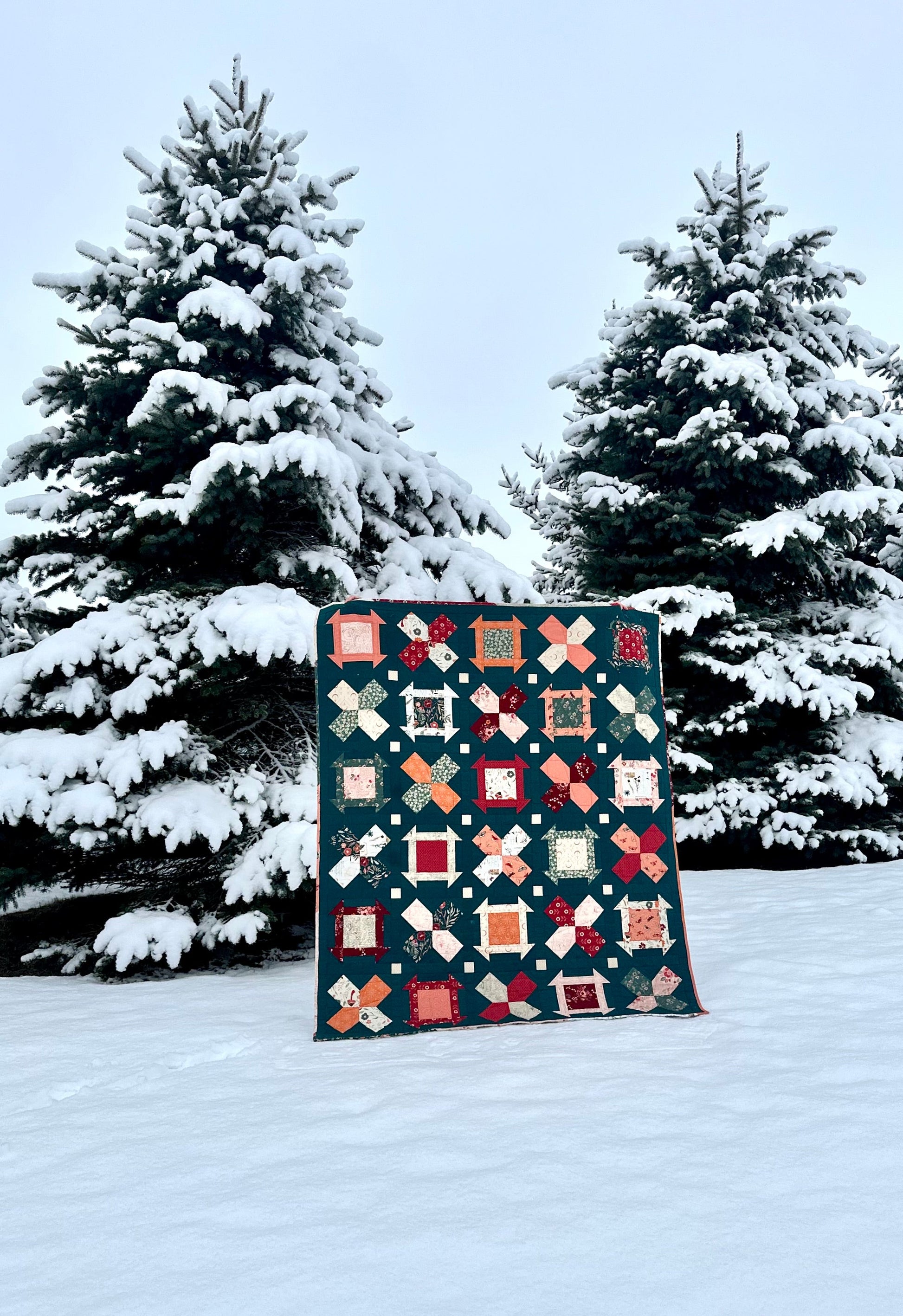 Colorful quilt featuring geometric patterns, set against a snowy landscape with evergreen trees.